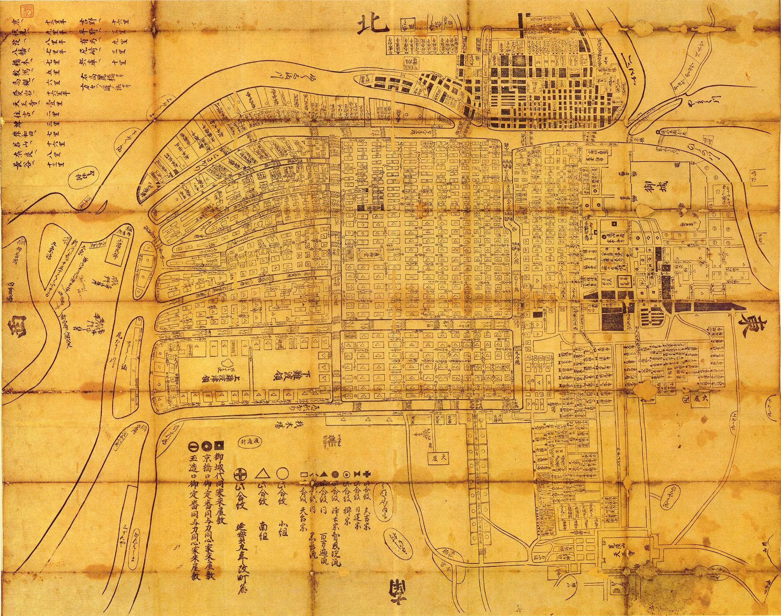 Line only, no colour, printed on paper now browned, the fold grid running through it. Every block is a small rectangle with its name set inside, and there are hundreds of them. A key of symbols — circles, triangles, crosses — runs along the foot, for the Buddhist sects and for the quarters of the castle warden's yoriki and dōshin. A table of distances to other towns sits in the upper left corner.<br><br>Almost none of the water is river. It is straight, bridged on both banks and named, because it was dug. North is at the top, the castle at the right. The fan of narrow strips at the upper left is reclaimed ground laid out in one go, and still the shape of Nishi-ku.